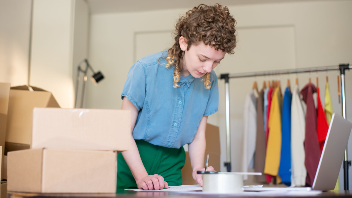 Online store owner at her desk with a laptop, shipping boxes and a clothing rack behind her